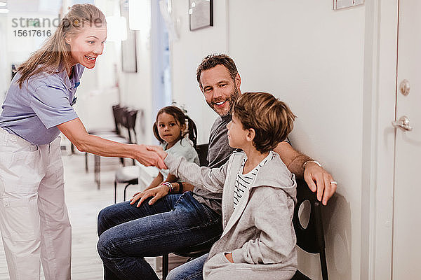 Lächelnde Kinderärztin beim Händeschütteln mit einem Jungen  der mit seiner Familie im Krankenhauskorridor sitzt