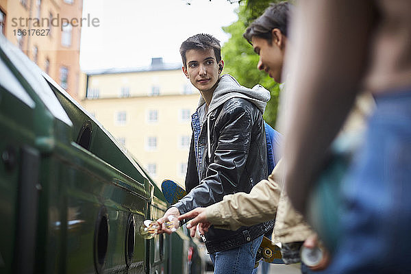 Teenager  der einen Freund ansieht  während er Abfälle in den Mülleimer einer Recycling-Station wirft