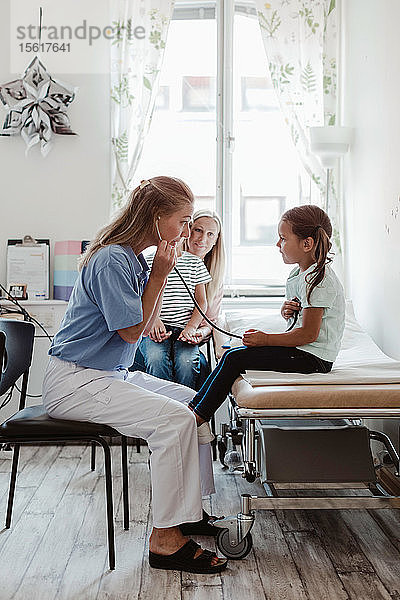 Kinderarzt hört den Herzschlag des Mädchens durch ein Stethoskop ab  während die Mutter mit dem Jungen im Hintergrund in der Klinik sitzt