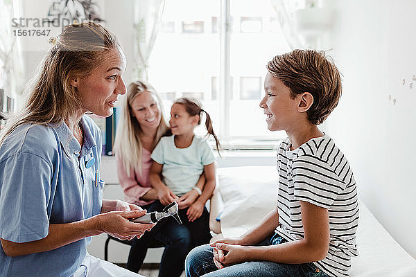 Lächelnde Kinderärztin mit Otoskop im Gespräch mit dem Jungen  während die Familie im Hintergrund in der medizinischen Klinik sitzt
