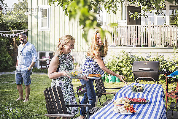 Freundinnen arrangieren Essen auf dem Tisch für geselliges Beisammensein im Garten am Sommerwochenende