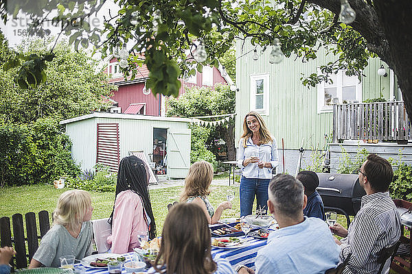 Glückliche Frau hält Rede  während sie während einer Gartenparty mit Freunden bei Tisch anstößt