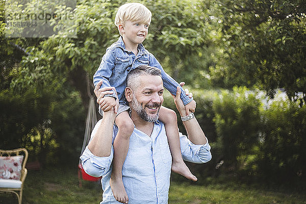 Lächelnder reifer Mann trägt seinen Sohn auf der Schulter  während er sich im Hinterhof vergnügt