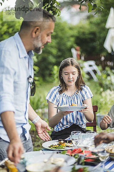 Mädchen hält Speiseteller  während sie mit der Familie im Garten am Tisch sitzt