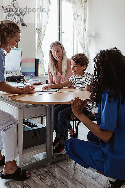 Arzt betrachtet Patient  der auf Papier zeigt  während Krankenschwester und Mutter im Krankenhaus am Schreibtisch sitzen
