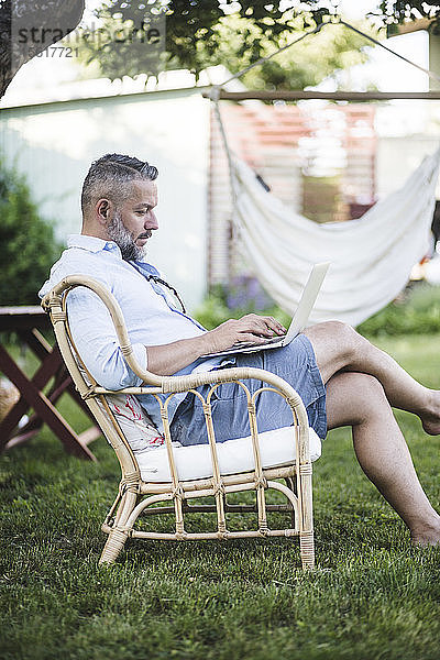 Seitenansicht eines reifen Mannes  der am Laptop arbeitet  während er auf einem Stuhl im Hinterhof sitzt