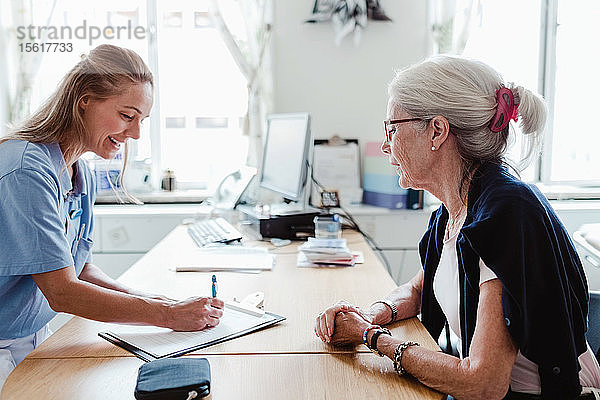 Lächelnder Arzt schreibt ein Rezept  während eine ältere Frau im Krankenhaus am Schreibtisch sitzt