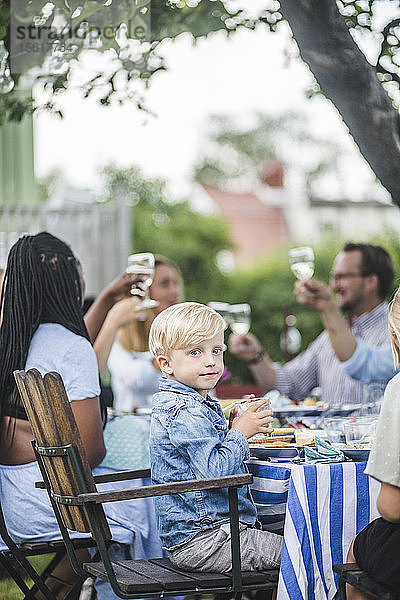 Junge trinkt  während er auf einem Stuhl bei einer Gartenparty mit Freunden und Familie sitzt