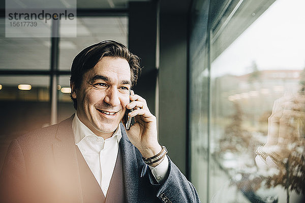 Mittelgroßer erwachsener Geschäftsmann spricht am Smartphone  während er im Büro am Fenster steht