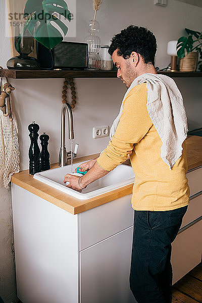 Seitenansicht eines mittelgroßen erwachsenen Mannes  der sein Geschirr an der Spüle in der Küche wäscht