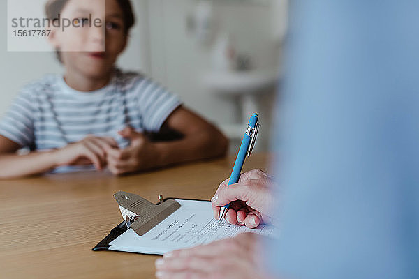 Geschnittene Hände eines Arztes  der ein Rezept schreibt  während ein Junge in der Klinik am Schreibtisch sitzt