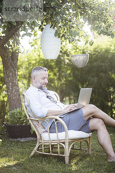 Reifer Mann tippt am Laptop  während er auf einem Holzstuhl im Hinterhof sitzt