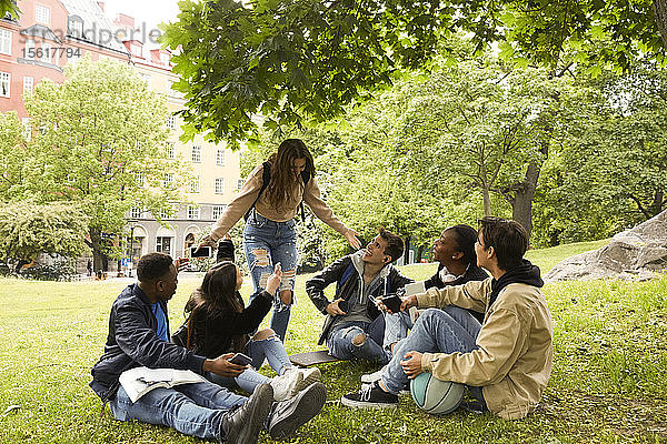 Glückliche Teenagerin gestikuliert  während sie bei Freunden steht  die im Park an Land sitzen