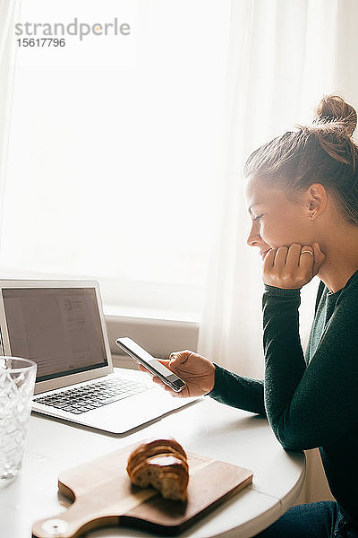 Mittelgroße erwachsene Frau  die ein Smartphone benutzt  während sie zu Hause mit einem Laptop auf einem Lebensmittelrezept surft