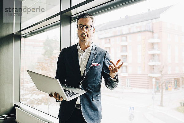 Reifer Geschäftsmann mit Kopfhörern  der mit seinem Laptop gegen ein Fenster im Firmenbüro spricht