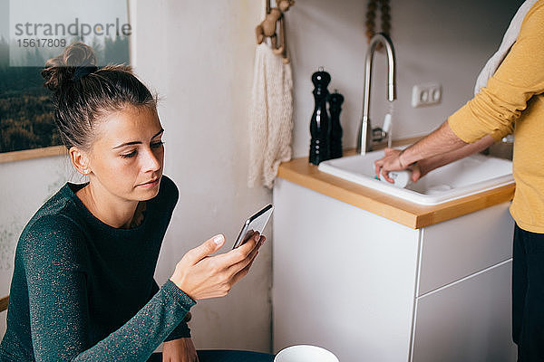 Mittlere erwachsene Frau  die ein Mobiltelefon benutzt  während ihr Freund zu Hause Utensilien wäscht