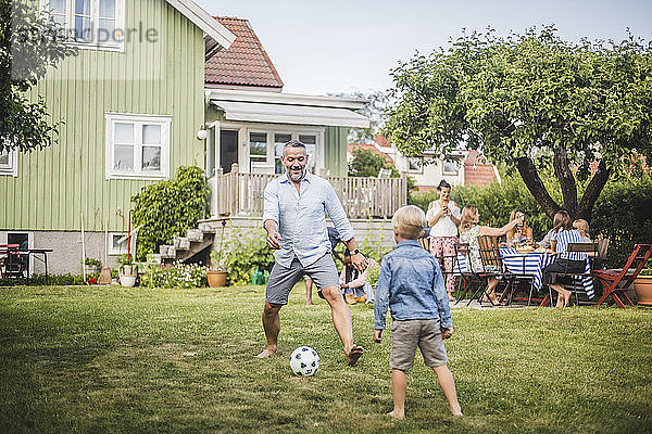 Vater spielt mit seinem Sohn Fussball  während Freunde sich am Tisch im Hinterhof amüsieren