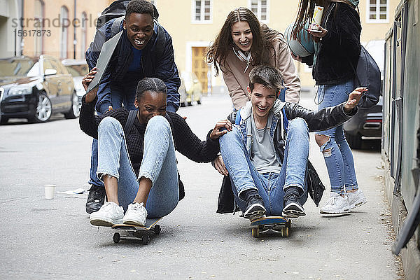 Fröhliche Freunde schubsen Teenager  die auf Skateboard sitzen