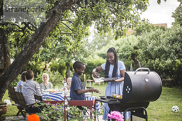 Lächelnde Mutter und Sohn bereiten während der Gartenparty gemeinsam Essen auf dem Grill zu