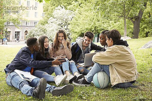 Lächelnder Teenager zeigt Freunden sein Smartphone  während er im Park sitzt
