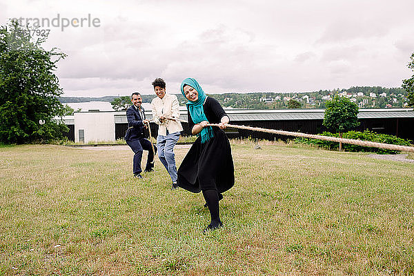 Lächelnde multiethnische Kollegen ziehen am Seil  während sie auf dem Rasen Tauziehen spielen