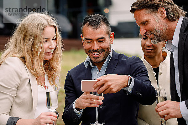Lächelnder Geschäftsmann zeigt männlichen und weiblichen Kollegen sein Handy  während er eine Sektflöte hält