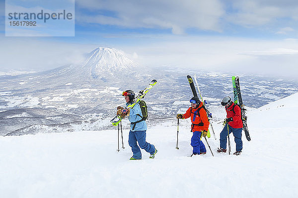 Mit dem Vulkan Yotei im Hintergrund wandern eine Skiläuferin und zwei Skiläufer zum Gipfel des Annupuri im Skigebiet Niseko United auf der japanischen Insel Hokkaido. Niseko United besteht aus vier Skigebieten auf dem Annupuri (1.308 m). Der 100 km südlich von Sapporo gelegene Niseko Annupuri ist Teil des Quasi-Nationalparks Niseko-Shakotan-Otaru Kaigan und ist der östlichste Park der Niseko-Vulkangruppe. Hokkaido  die nördliche Insel Japans  liegt geografisch ideal in der Bahn der beständigen Wettersysteme  die die kalte Luft aus Sibirien über das Japanische Meer bringen. Dies führt dazu  dass viele der Skigebiete mit Pulverschnee überhäuft werden  der für seine unglaubliche Trockenheit bekannt ist. In einigen der Skigebiete Hokkaidos fallen durchschnittlich 14-18 Meter Schnee pro Jahr. Niseko ist die Pulverschneehauptstadt der Welt und als solche das beliebteste internationale Skigebiet in Japan. Es bietet ein unvergessliches Erlebnis für Skifahrer und Snowboarder aller Niveaus. Der Berg Yotei im Hintergrund wird oft auch als der Berg Fuji von Hokkaido bezeichnet.