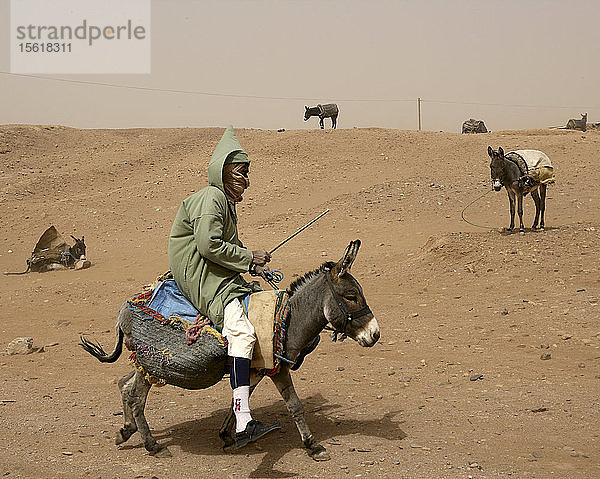Mann reitet auf einem Esel in einer staubigen Sandstraße in der Kasbah von M'hamid in Südmarokko