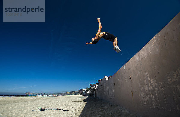 Ein athletischer Mann macht einen Rückwärtssalto von einer Mauer auf den Strand in San Diego  CA.
