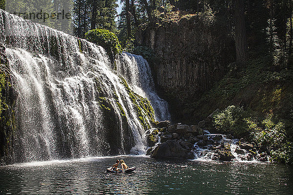 Entfernte Ansicht eines Paares in einem Floß  das auf dem Wasser in der Nähe eines Wasserfalls treibt  ï¾ McCloudï¾ River  Kalifornien  USA