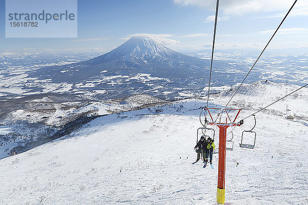 Vor dem Hintergrund des Vulkans Yotei sitzen eine Skifahrerin und ein Skifahrer in einem Sessellift  der sie zum Gipfel des Annupuri im Skigebiet Niseko United auf der japanischen Insel Hokkaido bringt. Niseko United besteht aus vier Skigebieten auf dem Annupuri (1.308 m). Der 100 km südlich von Sapporo gelegene Niseko Annupuri ist Teil des Quasi-Nationalparks Niseko-Shakotan-Otaru Kaigan und ist der östlichste Park der Niseko-Vulkangruppe. Hokkaido  die nördliche Insel Japans  liegt geografisch ideal in der Bahn der beständigen Wettersysteme  die die kalte Luft aus Sibirien über das Japanische Meer bringen. Dies führt dazu  dass viele der Skigebiete mit Pulverschnee überhäuft werden  der für seine unglaubliche Trockenheit bekannt ist. In einigen der Skigebiete Hokkaidos fallen durchschnittlich 14-18 Meter Schnee pro Jahr. Niseko ist die Pulverschneehauptstadt der Welt und als solche das beliebteste internationale Skigebiet in Japan. Es bietet ein unvergessliches Erlebnis für Skifahrer und Snowboarder aller Niveaus. Der Berg Yotei im Hintergrund wird oft auch als der Berg Fuji von Hokkaido bezeichnet.