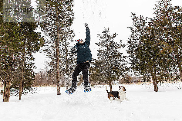 Ein Mann in Schneeschuhen springt im Winter mit seinem Beagle durch den Schnee.