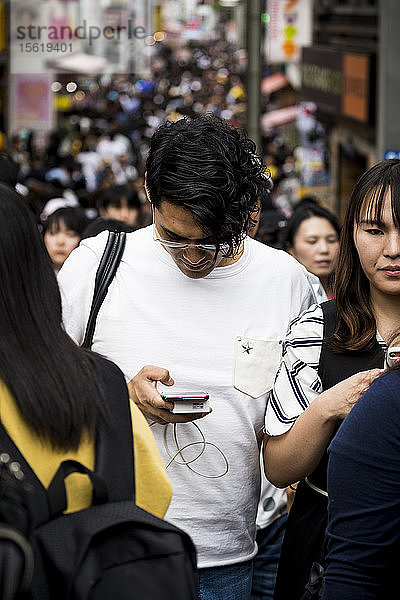 Junger Mann  der inmitten einer belebten Straße auf sein Smartphone schaut  Harijuku  Tokio  Japan