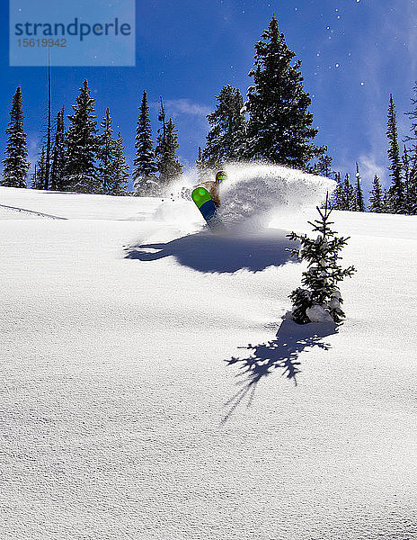 Ein Mann kommt an einem sonnigen Wintertag mit seinem Snowboard aus einer Pulverschneewolke heraus.