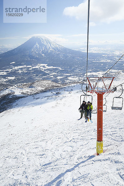 Vor dem Hintergrund des Vulkans Yotei sitzen eine Skifahrerin und ein Skifahrer in einem Sessellift  der sie zum Gipfel des Annupuri im Skigebiet Niseko United auf der japanischen Insel Hokkaido bringt. Niseko United besteht aus vier Skigebieten auf dem Annupuri (1.308 m). Der 100 km südlich von Sapporo gelegene Niseko Annupuri ist Teil des Quasi-Nationalparks Niseko-Shakotan-Otaru Kaigan und ist der östlichste Park der Niseko-Vulkangruppe. Hokkaido  die nördliche Insel Japans  liegt geografisch ideal in der Bahn der beständigen Wettersysteme  die die kalte Luft aus Sibirien über das Japanische Meer bringen. Dies führt dazu  dass viele der Skigebiete mit Pulverschnee überhäuft werden  der für seine unglaubliche Trockenheit bekannt ist. In einigen der Skigebiete Hokkaidos fallen durchschnittlich 14-18 Meter Schnee pro Jahr. Niseko ist die Pulverschneehauptstadt der Welt und als solche das beliebteste internationale Skigebiet in Japan. Es bietet ein unvergessliches Erlebnis für Skifahrer und Snowboarder aller Niveaus. Der Berg Yotei im Hintergrund wird oft auch als der Berg Fuji von Hokkaido bezeichnet.