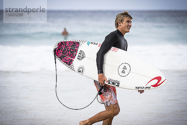 Seitenansicht eines einzelnen lächelnden männlichen Surfers  der ein Surfbrett am Strand entlang trägt
