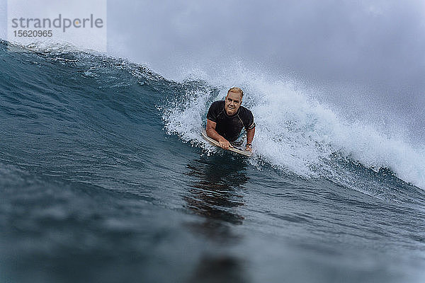 Männlicher Surfer schaut in die Kamera  während er auf dem Surfbrett liegt und eine Welle reitet  Male  Malediven