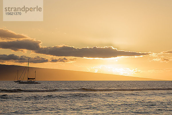 Ein Segelboot bei Sonnenuntergang mit einer Insel in der Ferne vor der Küste von Maui.