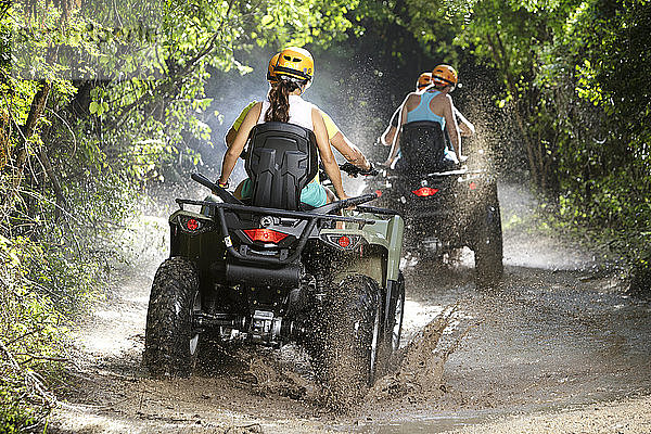 Rückansicht von Paaren  die mit Quad-Bikes durch Pfützen auf unbefestigten Straßen im Emotions Native Park  Quintana Roo  Mexiko fahren
