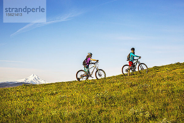 Zwei junge Frauen schieben ihre Mountainbikes auf einem einspurigen Weg durch eine offene Wiese.