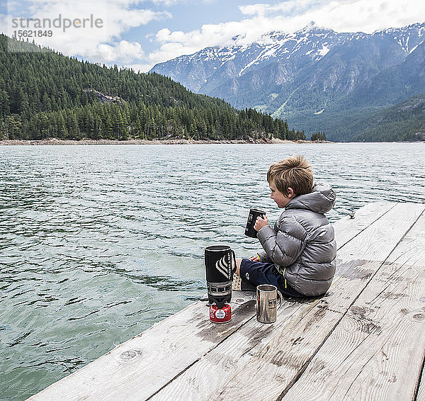 Ein Junge sitzt auf einem Steg und trinkt Tee am Ross Lake im North Cascades National Park in Washington.