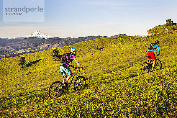 Zwei junge Frauen fahren mit ihren Mountainbikes auf einem einspurigen Weg durch eine offene Wiese mit einem Vulkan in der Ferne.
