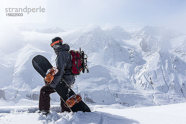 Ein Backcountry-Snowboarder mit seinem Board steht auf einem Bergrücken und überblickt die entfernten Berge