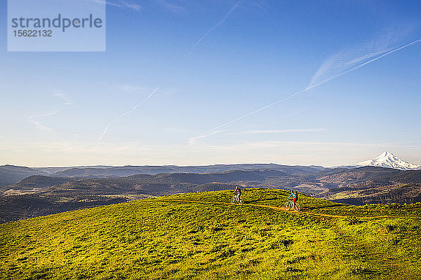 Zwei junge Frauen fahren mit dem Mountainbike auf einem einspurigen Weg durch grünes Gras in der frühen Morgensonne.