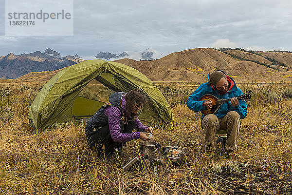 Eine Camperin bereitet das Frühstück auf einem Campingkocher zu  während ein Camper vor seinem Zelt sitzt und Mandoline spielt  Jackson  Wyoming  USA