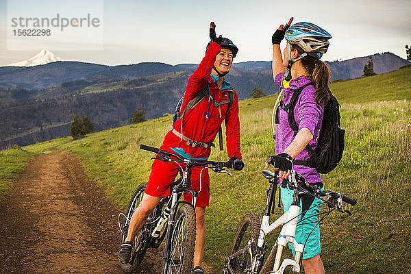 Zwei junge Frauen auf Mountainbikes geben sich High-Five  während sie sich auf einer Wiese unter dem frühen Morgenhimmel mit dem Vulkan in der Ferne ausruhen.