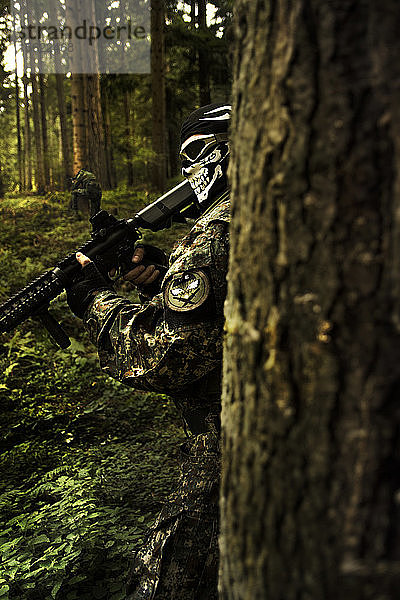 Maskierter Soldat mit einem Gewehr hinter einem Baum versteckt. Im Hintergrund hockt ein weiterer Soldat  ebenfalls hinter einem Baum. Fotografiert während eines Airsoft-Spiels auf dem Berg Goc  Zentralserbien.