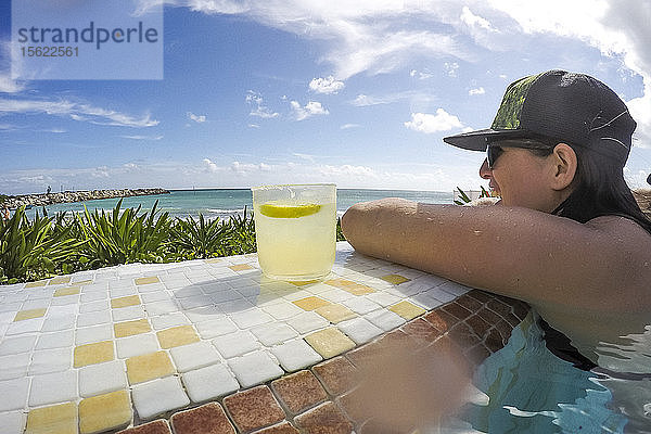 Frau entspannt sich am Rande eines Swimmingpools am Meer mit einem Cocktailempfang