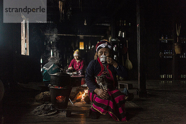 Porträt einer älteren Frau  die hinter einem Lagerfeuer sitzt  eine Katze auf dem Schoß hat und aus einer Tasse trinkt  Myanmar  Shan  Myanmar