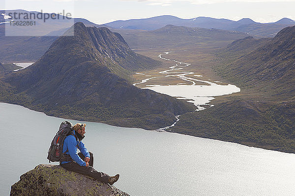 Ein Wanderer genießt die Aussicht auf Fjorde und Berge  während er auf einem Felsen des berühmten Besseggen-Kamms in Norwegen sitzt. Diese eintägige Überquerung im Jotunheimen-Nationalpark ist die beliebteste Wanderung in Norwegen.